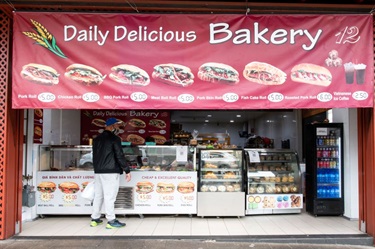 Shop front of Daily Delicious Bakery at Dutton Plaza featuring a large banner with menu of bánh mì sandwiches and other various baked foods visible through the glass counter