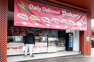 Shop front of Daily Delicious Bakery at Dutton Plaza featuring a large banner with menu of bánh mì sandwiches and other various baked foods visible through the glass counter