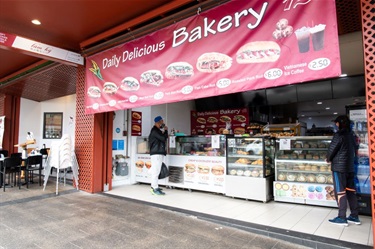Shop front of Daily Delicious Bakery at Dutton Plaza featuring a large banner with menu of bánh mì sandwiches and other various baked foods visible through the glass counter