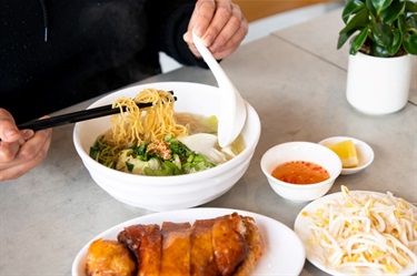 Close up of person's hands using chopsticks and spoon to pull noodles from a Vietnamese egg noodle soup while surrounded by sliced crispy skin chicken and various condiments at Lam Ky Noodle House in Dutton Plaza