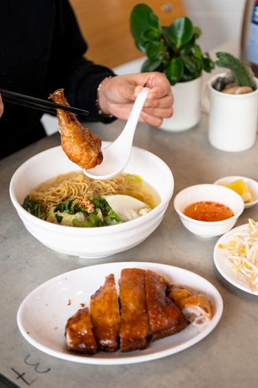 Close up of person's hands using chopsticks and spoon to hold a crispy skin chicken drumstick above a Vietnamese egg noodle soup, sliced crispy skin chicken and various condiments at Lam Ky Noodle House in Dutton Plaza