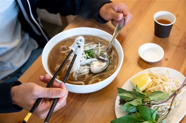 Closeup of a person's hands using a spoon and chopsticks to eat pho noodle soup at Lucky Cat Vietnamese Restaurant in Dutton Plaza